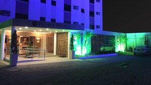 L’entrée d’un hôtel, avec une enseigne indiquant « PALACE HOTEL », un espace extérieur couvert doté d’une rambarde et un parking avec des voitures.