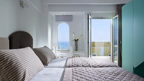 Habitación de hotel moderna con una cama grande, un área de descanso con vista al mar y un balcón con puerta de vidrio.