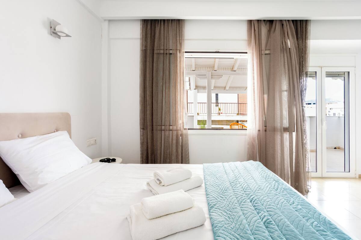 A hotel room with a bed, a window with curtains, a view of the outside, and a door leading to a balcony.
