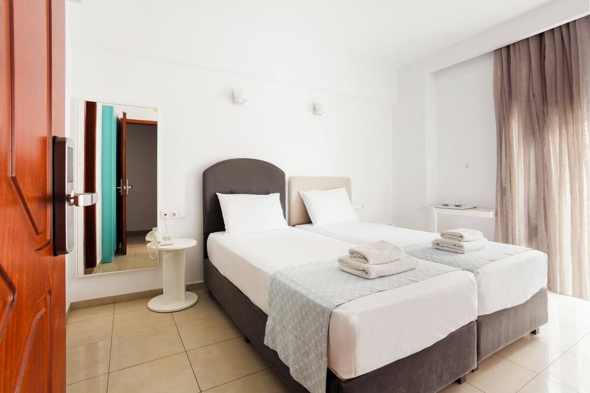 A hotel room with a bed, bedside tables, a mirror, a wardrobe, and a window with curtains.