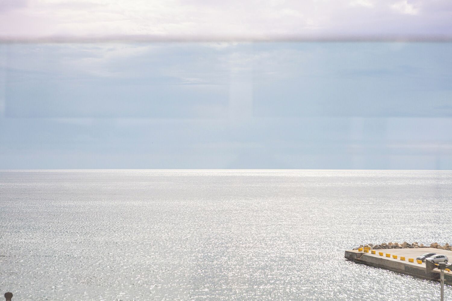 Uitzicht op de kust, met heldere lucht, kalme zee en een steiger met een auto en gele paaltjes.