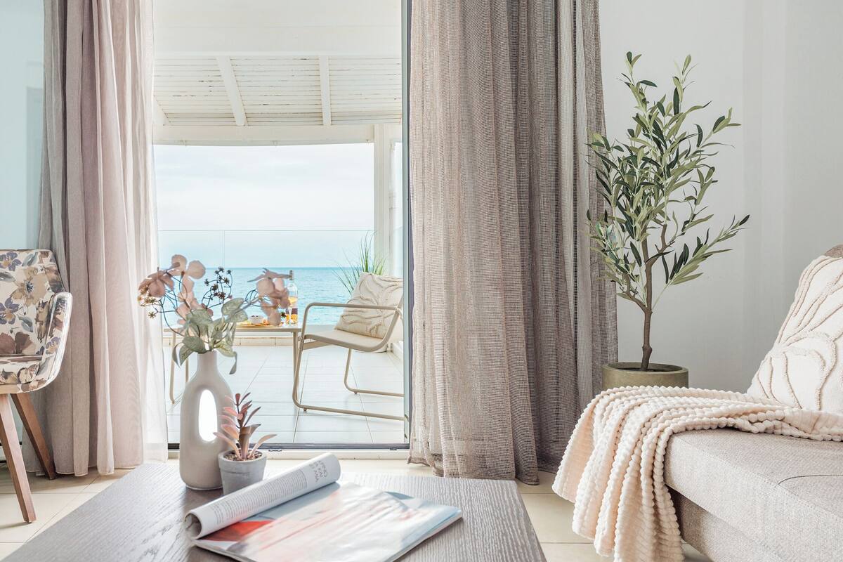 A room with a view of the sea, a sofa, a chair, a table with a plant, and a magazine.