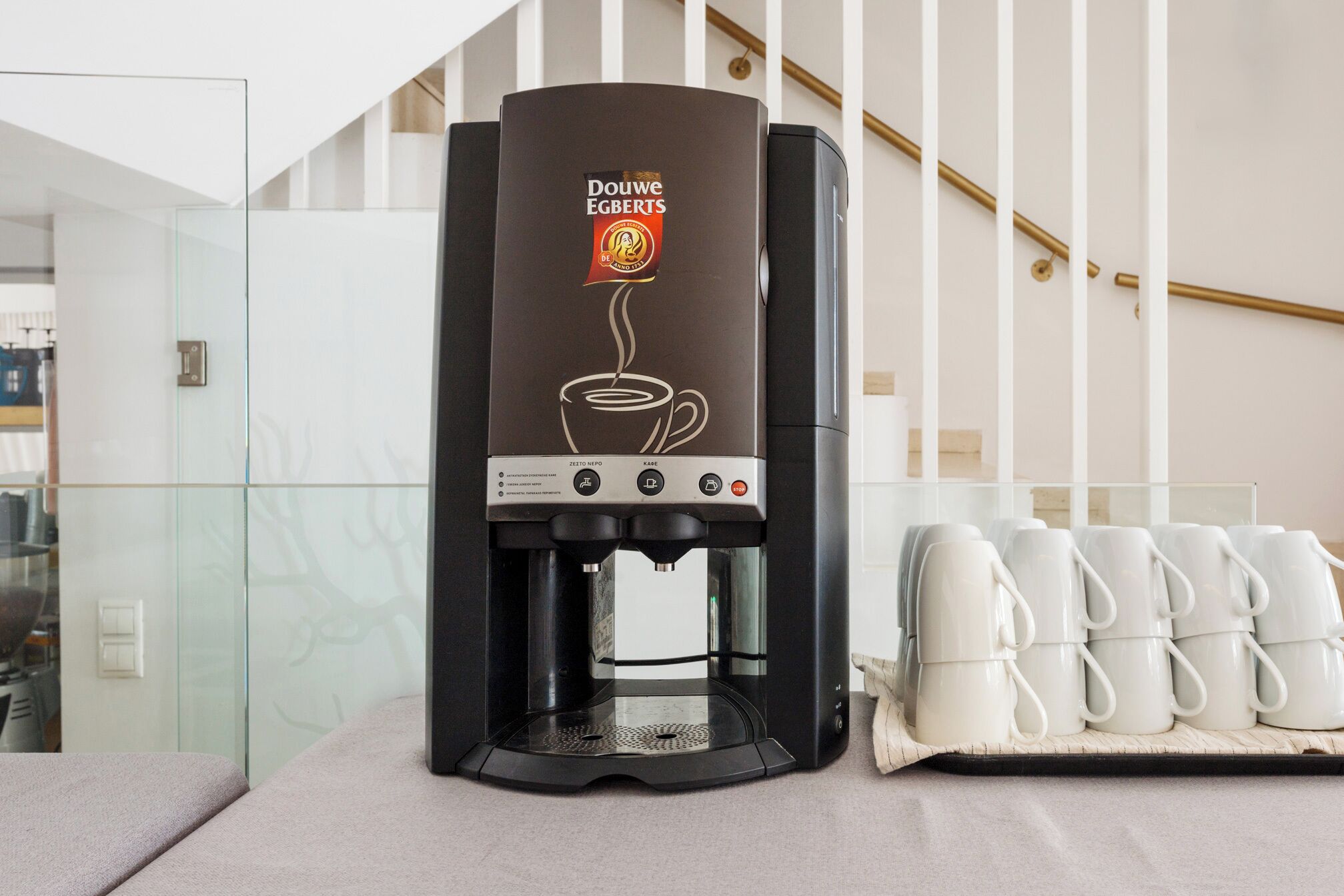 Una máquina de café Douwe Egberts con depósito de agua de vidrio, panel de control y bandeja con tazas blancas.