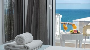 Habitación de hotel con vista al mar, una toalla blanca doblada con cuidado sobre la cama y una mesa con frutas y bebidas.