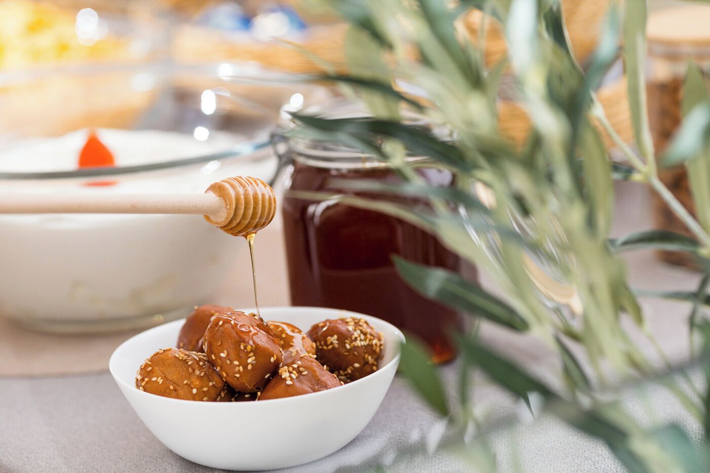 A bowl of sesame-seed-covered sweets with honey being drizzled on them, a jar of honey, and a candle in the background.