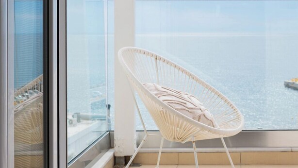 Un balcón con una silla de mimbre blanca, barandilla de vidrio y vista al mar.