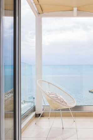 Un balcón con una silla de mimbre blanca, barandilla de vidrio y vista al mar.