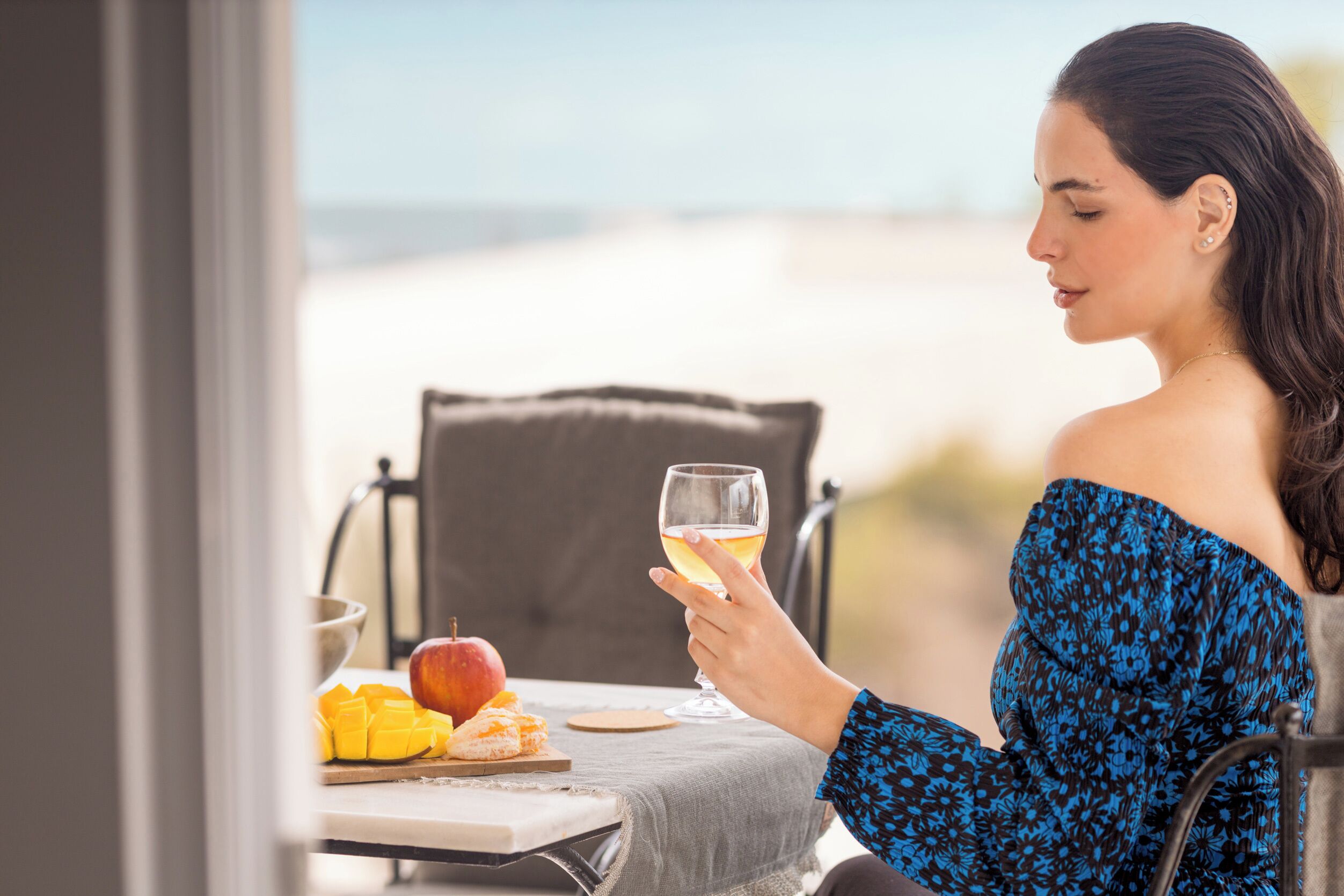 Una persona sentada a una mesa con una copa de vino, una manzana y fruta cortada, con vista a una playa.