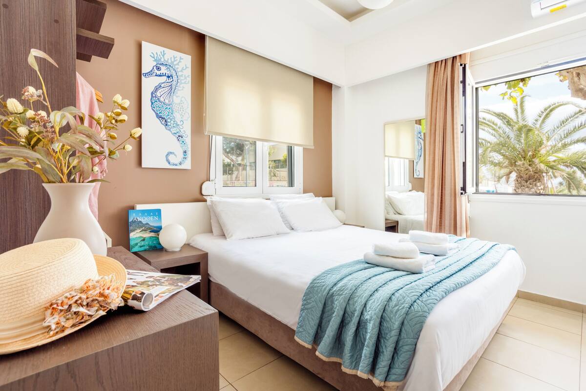 A hotel room with a bed, a nightstand, a vase with dried flowers, a book, and a window with a view of palm trees.