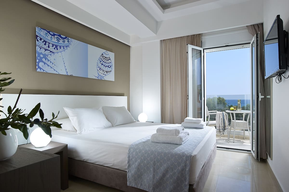 A modern hotel room with a large bed, a bedside table with a lamp, a balcony with a table and chairs, and a TV mounted on the wall.