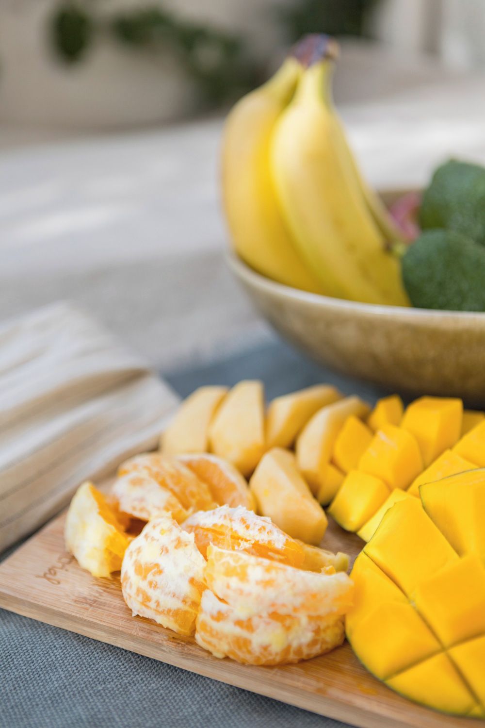 Una tabla de madera con mango en rodajas y segmentos de naranja, y un tazón con plátanos y hojas verdes al fondo.