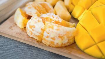 Una tabla de madera con mango en rodajas y segmentos de naranja, y un tazón con plátanos y hojas verdes al fondo.
