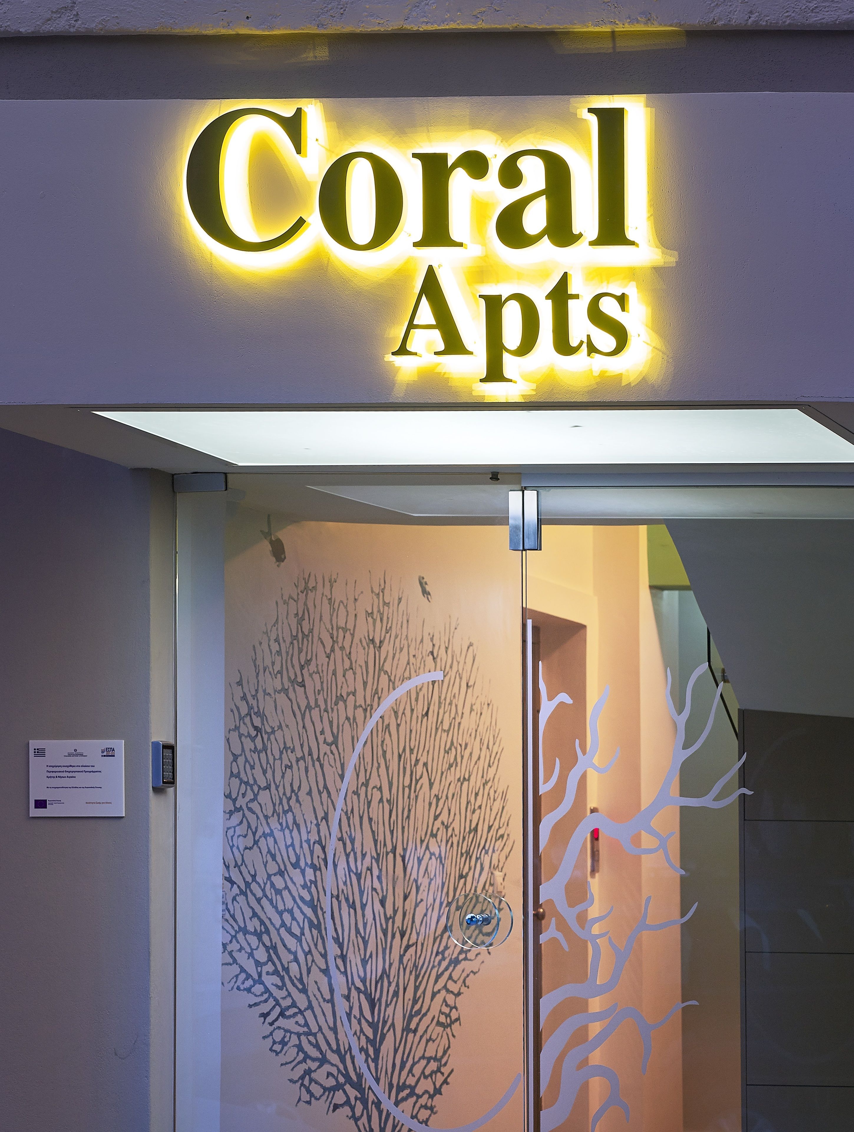L’entrée d’un immeuble, avec une enseigne indiquant « Coral Apts » et une porte décorative en verre ornée d’un motif représentant un arbre.
