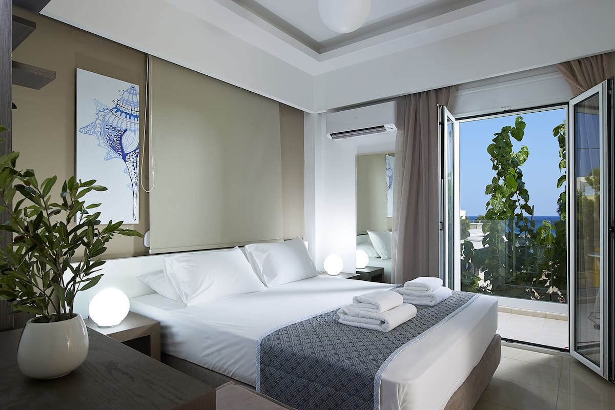 A modern hotel room with a large bed, a potted plant, a bedside table, and a view of the sea through a sliding glass door.