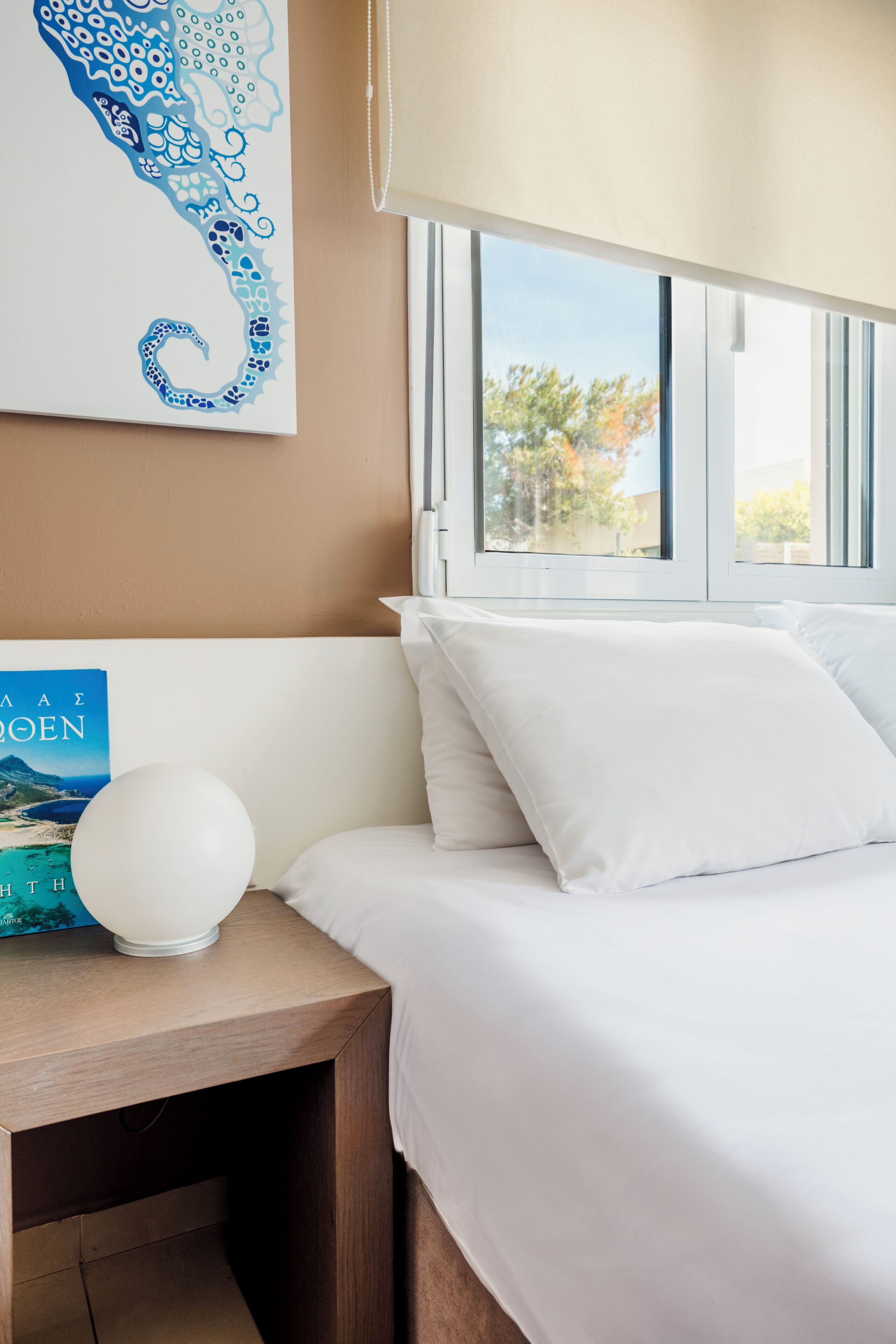 A bedroom with a bed, white pillows, a bedside table with a book and a lamp, a window with a view of trees, and a wall with a blue and white artwork.