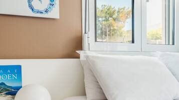 A bedroom with a bed, white pillows, a bedside table with a book and a lamp, a window with a view of trees, and a wall with a blue and white artwork.