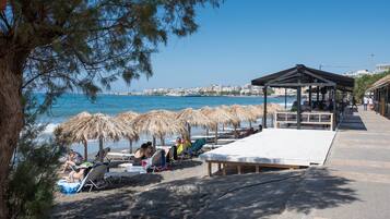 A beach with sun loungers and umbrellas, a wooden deck, and a view of the sea and buildings in the distance.