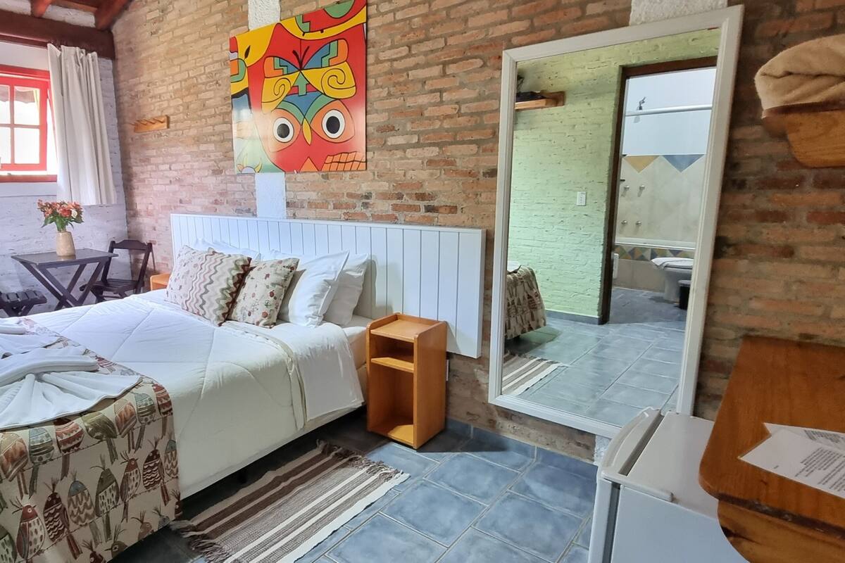 A cozy bedroom with a brick wall, a fireplace, a bed with patterned bedding, a wooden nightstand, and a large mirror.