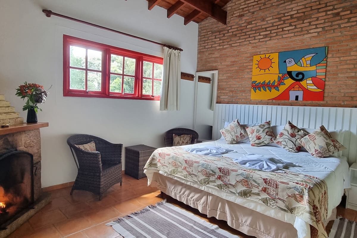 A cozy bedroom with a fireplace, a bed with floral bedding, a wicker chair, and a colorful painting on the wall.