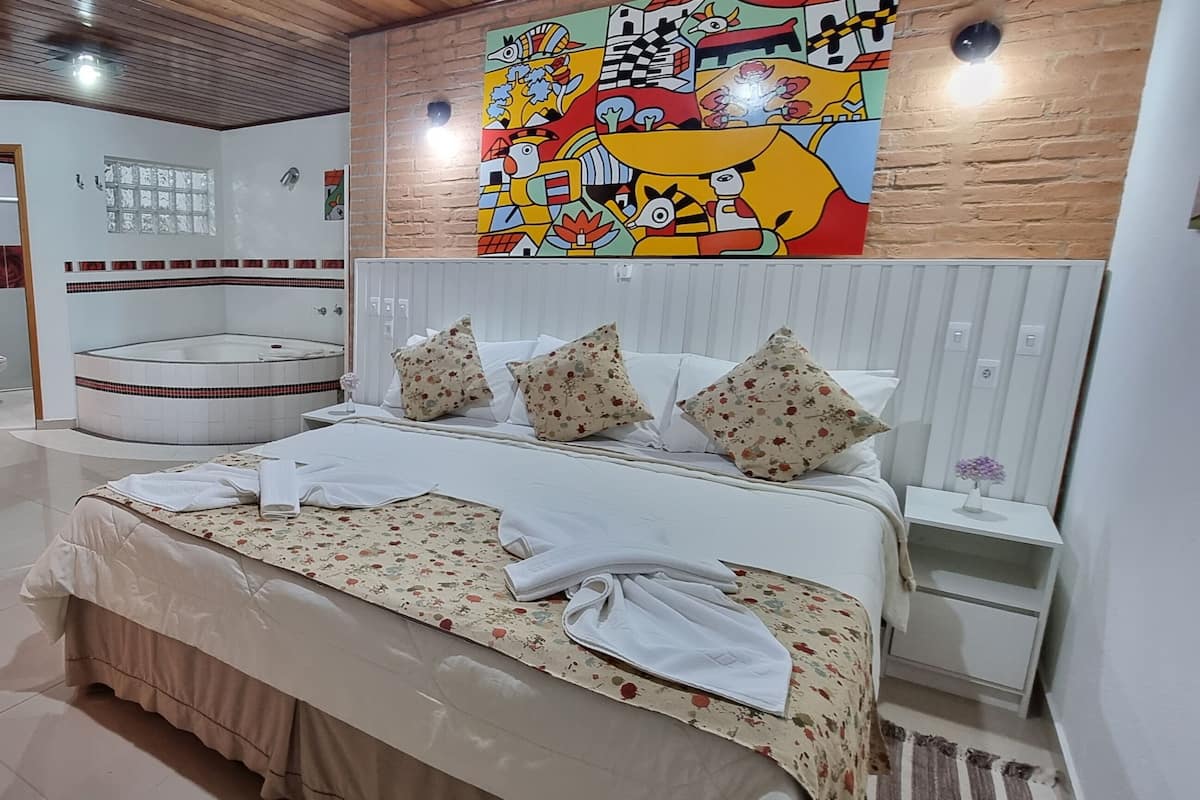 A modern bedroom with a large bed, a small table, and a chair. There is a colorful abstract painting on the wall and a tiled fireplace.