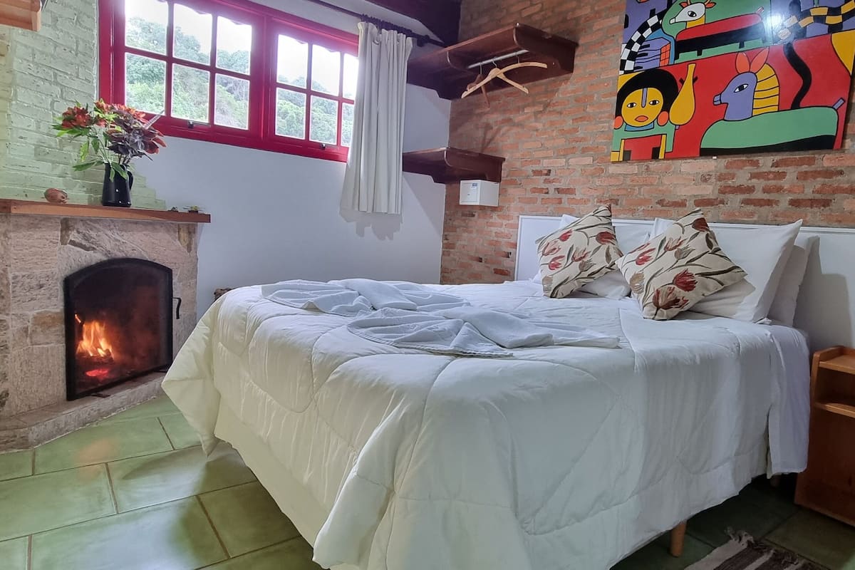 A room with a brick wall, a fireplace, a bed with floral pillows, and a colorful painting.