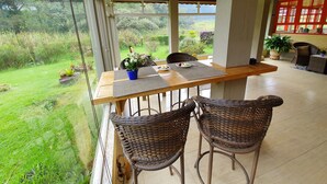 Una mesa de madera con dos sillas de mimbre, una planta en maceta y vistas a un jardín exuberante a través de ventanas amplias.