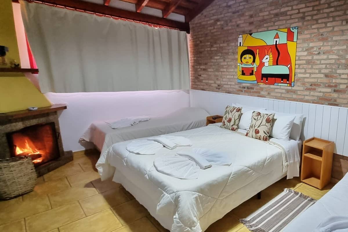 A cozy room with a bed, a nightstand, a wooden side table, a fireplace, and a colorful painting on the brick wall.