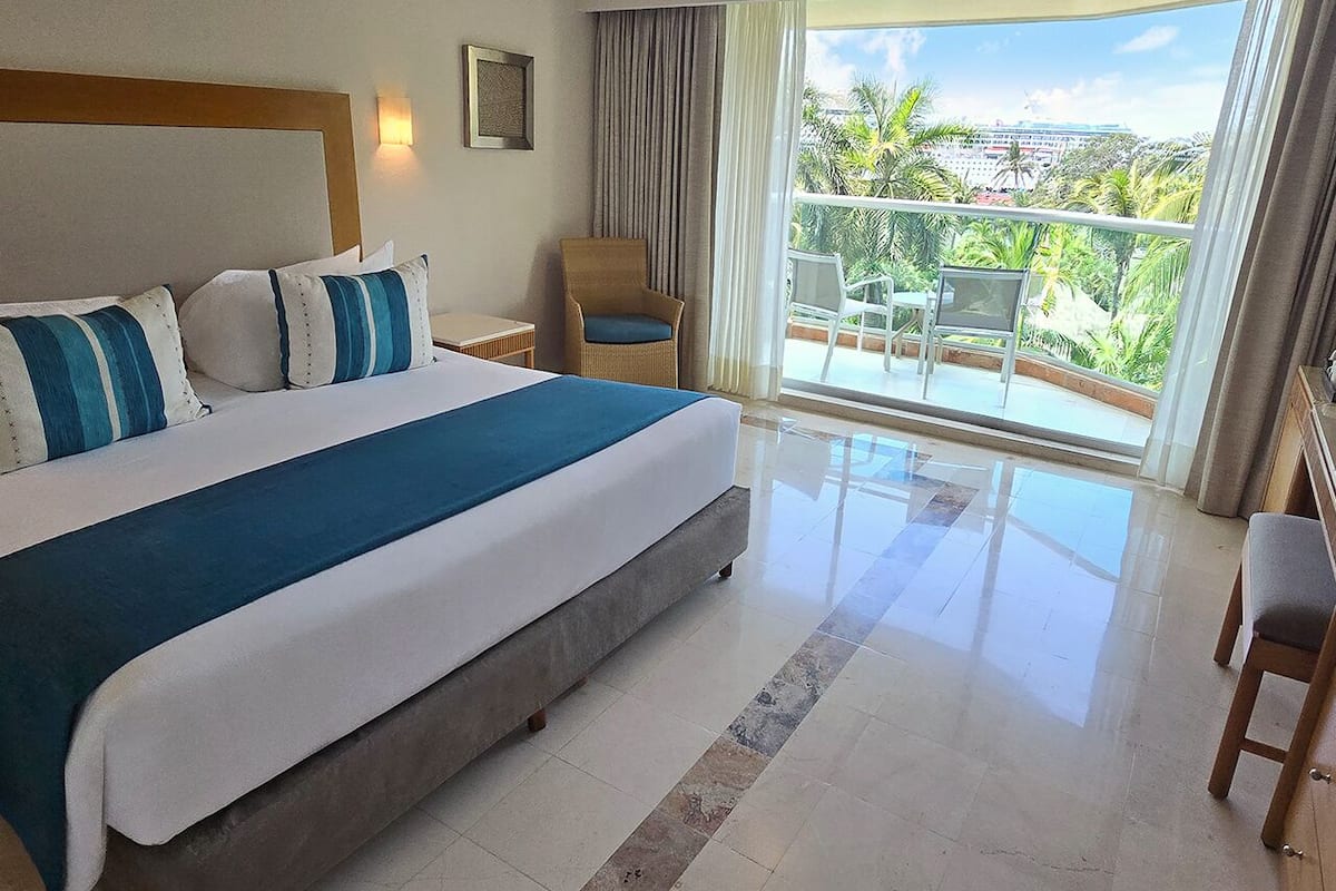 A hotel room with a large bed, a chair, a small table, and a balcony with a view of palm trees and a building.