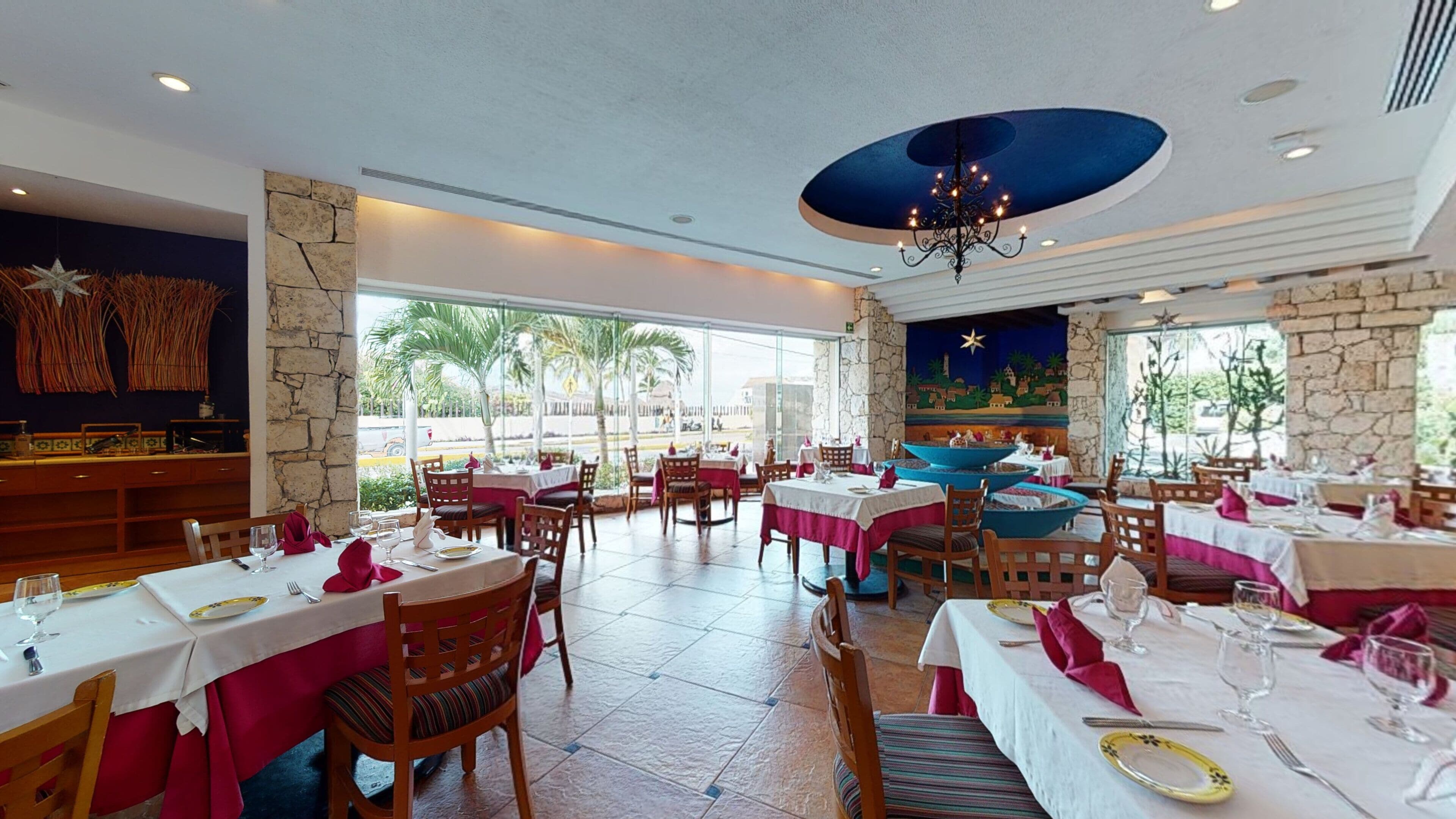 Un restaurant doté de nombreuses tables, de chaises et d’une grande fenêtre offrant une vue sur les palmiers.