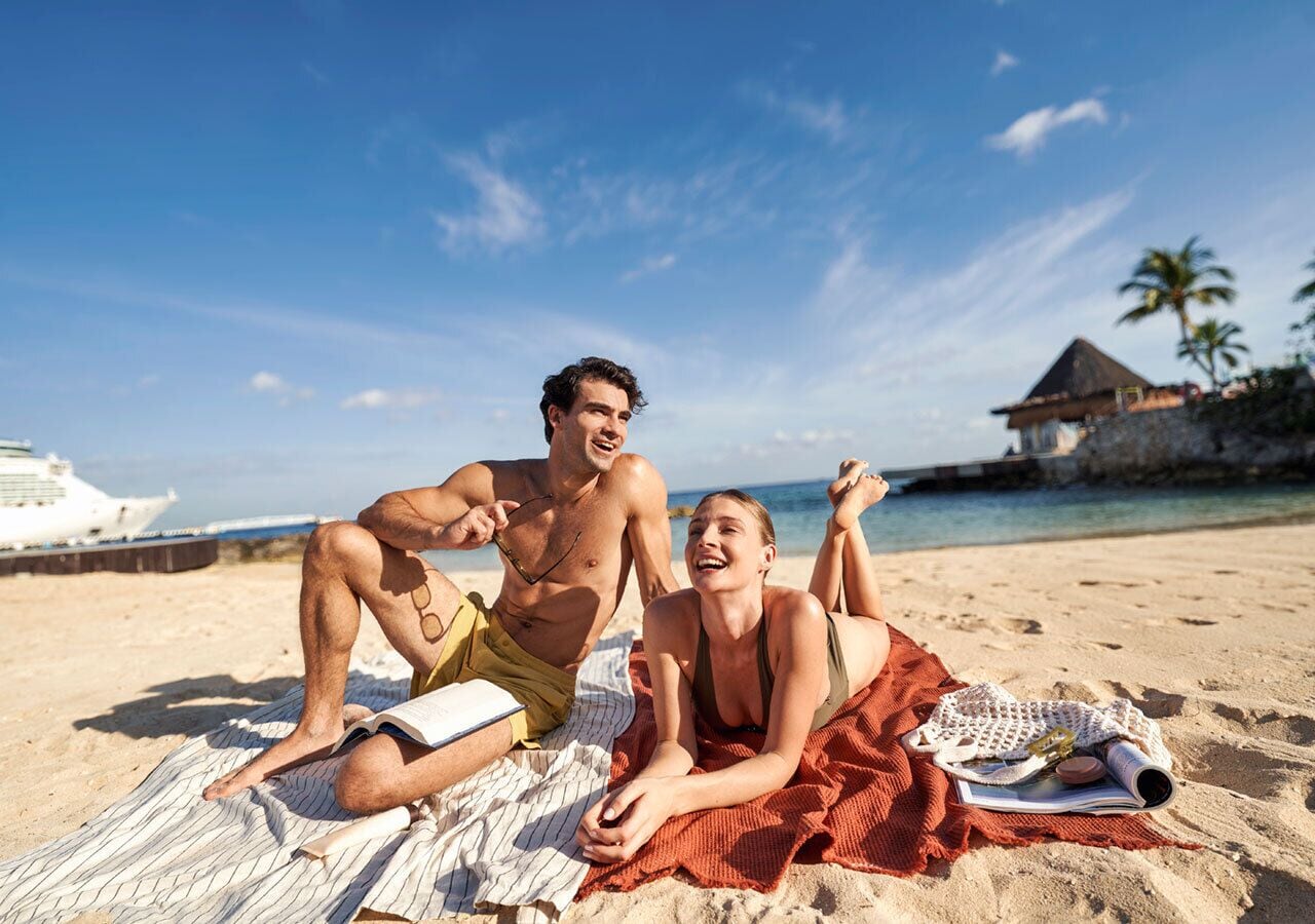Una pareja descansando sobre una toalla en la playa, con un bote y una palmera al fondo.