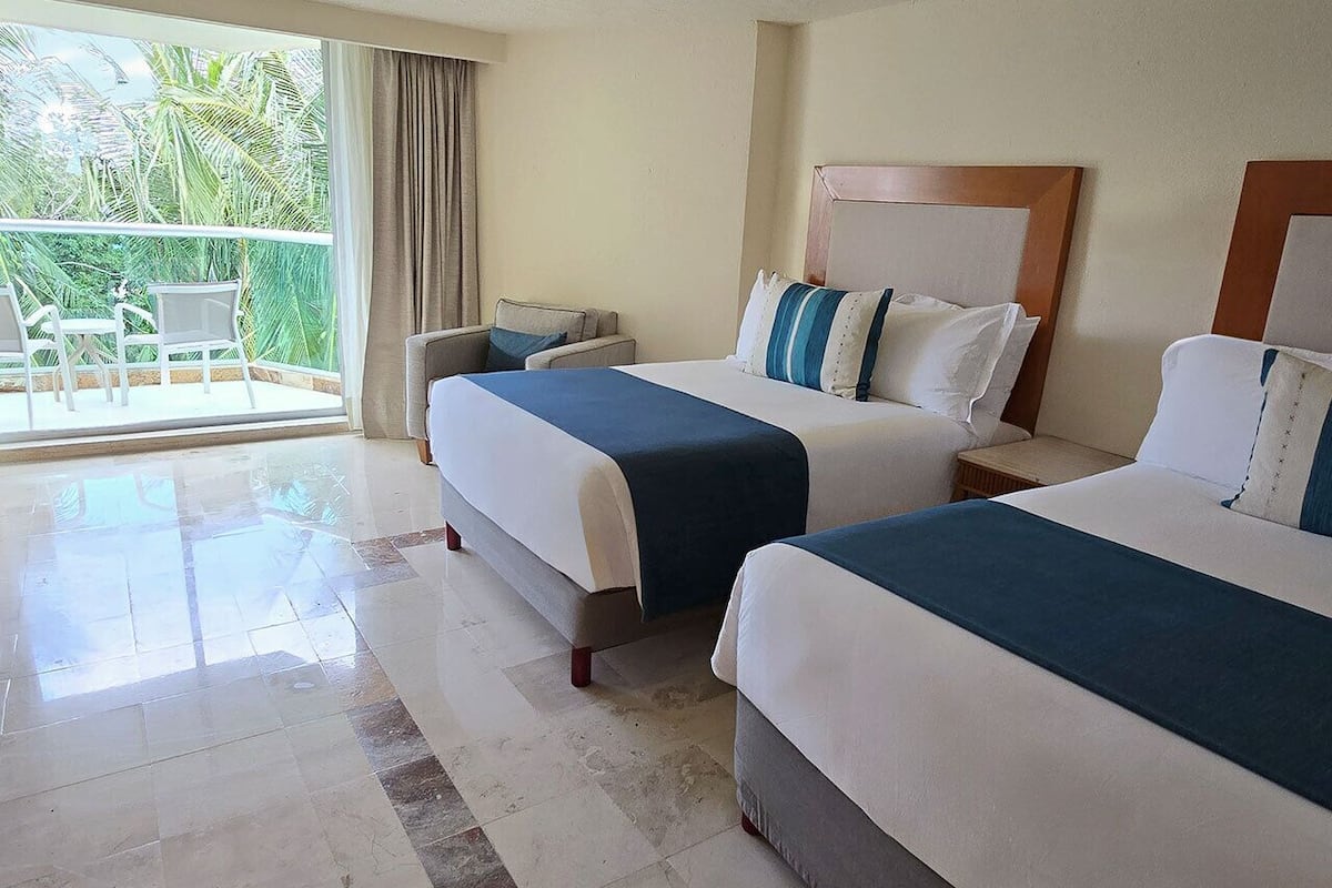 A hotel room with two beds, a balcony with chairs, and a view of palm trees.