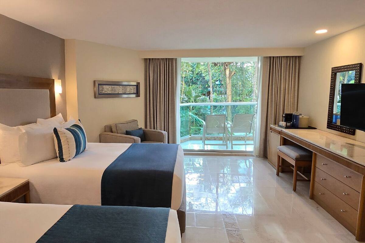A hotel room with two beds, a desk, a chair, a TV, and a balcony with a view of greenery.