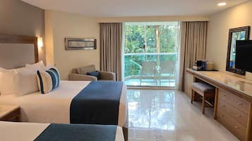 A hotel room with two beds, a desk, a chair, a TV, and a balcony with a view of greenery.