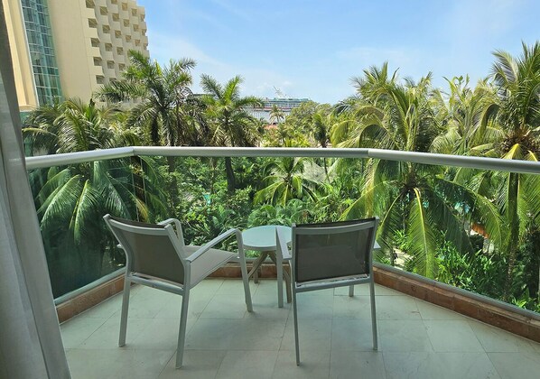A balcony with two chairs and a table, overlooking a lush garden and a building with a unique facade.
