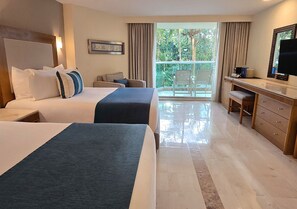 A hotel room with two beds, a desk, a chair, and a large window with a view of greenery.