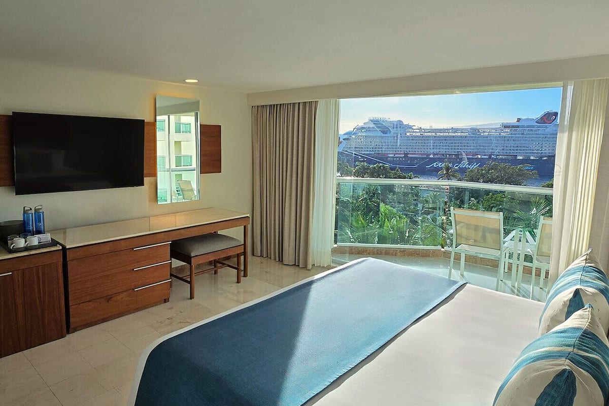 A hotel room with a bed, a desk, a TV, and a balcony with a view of a cruise ship.