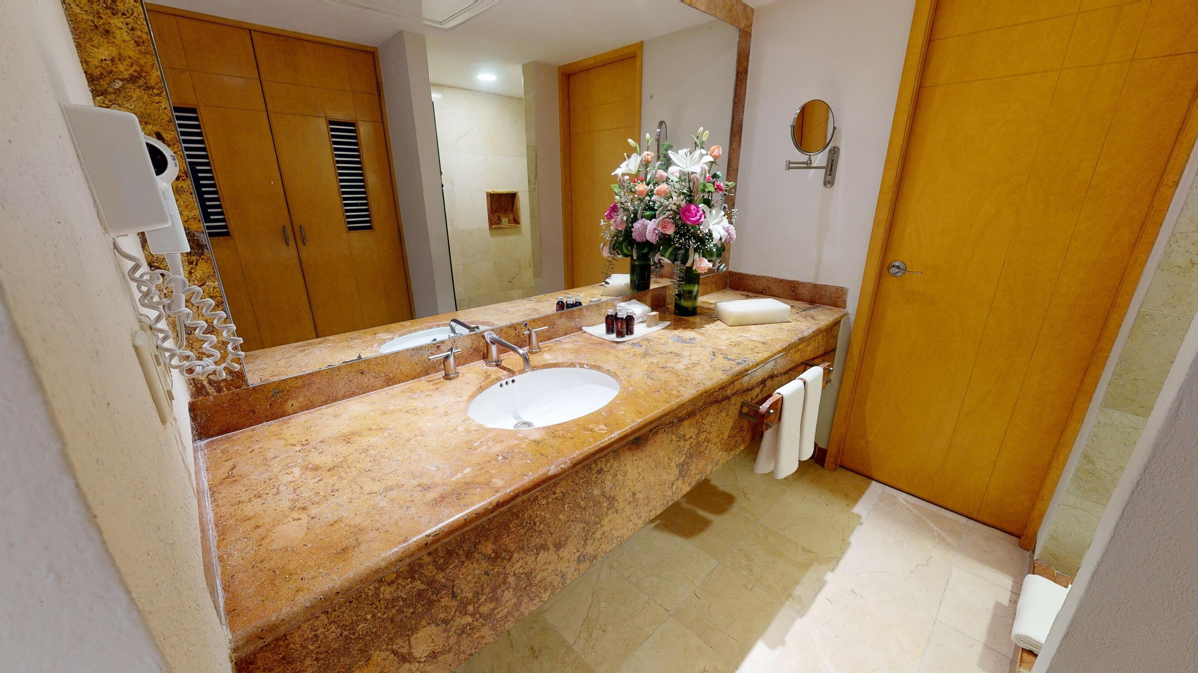Une salle de bain avec un lavabo en marbre, un grand miroir, une porte en bois et un vase de fleurs posé sur le comptoir.