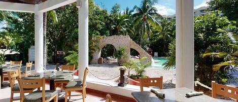 Un área de estar al aire libre con mesas y sillas de madera, un arco de piedra y una piscina al fondo.