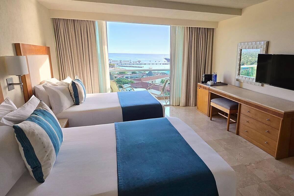 A hotel room with two beds, a desk, a television, and a view of the ocean.