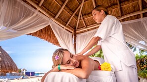 A person receiving a massage in a covered outdoor area with a thatched roof and white drapes.