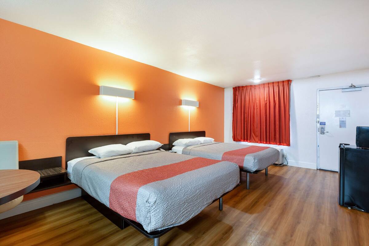A hotel room with two beds, a desk, and a chair. There are two wall-mounted lights and a door with a lock.