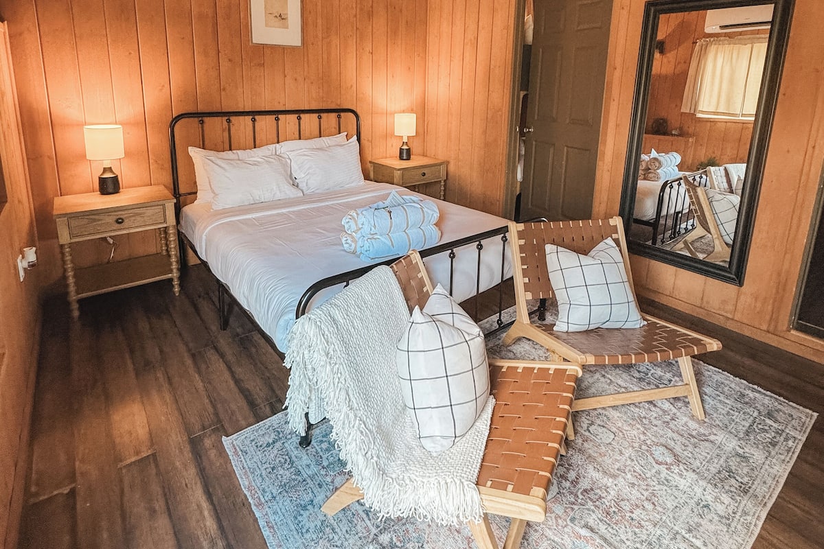 A wooden-paneled room with a bed, two bedside tables, a ceiling fan, a mirror, and a patterned rug.