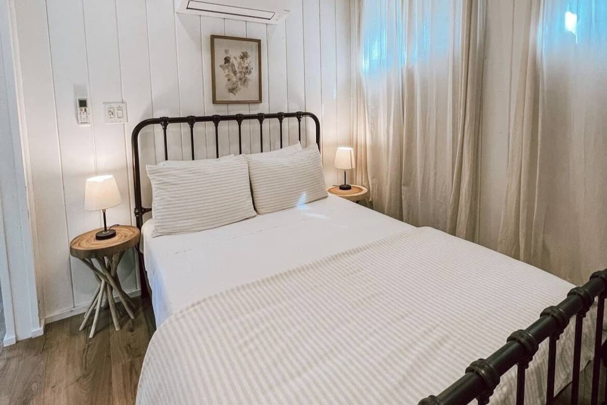 A bedroom with a wooden ceiling, a bed with white bedding, two bedside tables with lamps, and a wall-mounted air conditioner.
