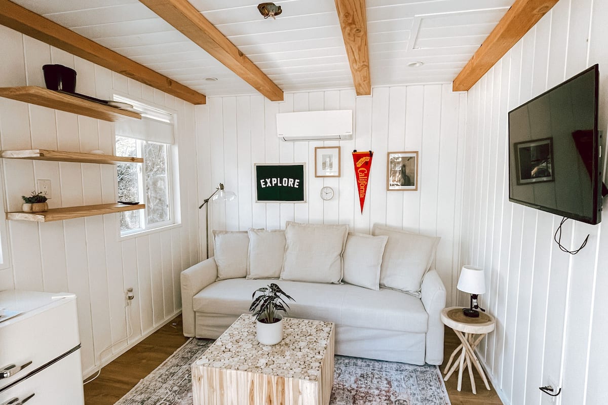 A cozy living room with a sofa, coffee table, and a television. There are wooden beams on the ceiling, a window with blinds, and a wall-mounted shelf.