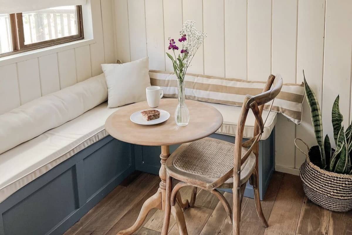 A cozy corner with a wooden bench, a round table with a cup and a slice of cake, and a vase with flowers.