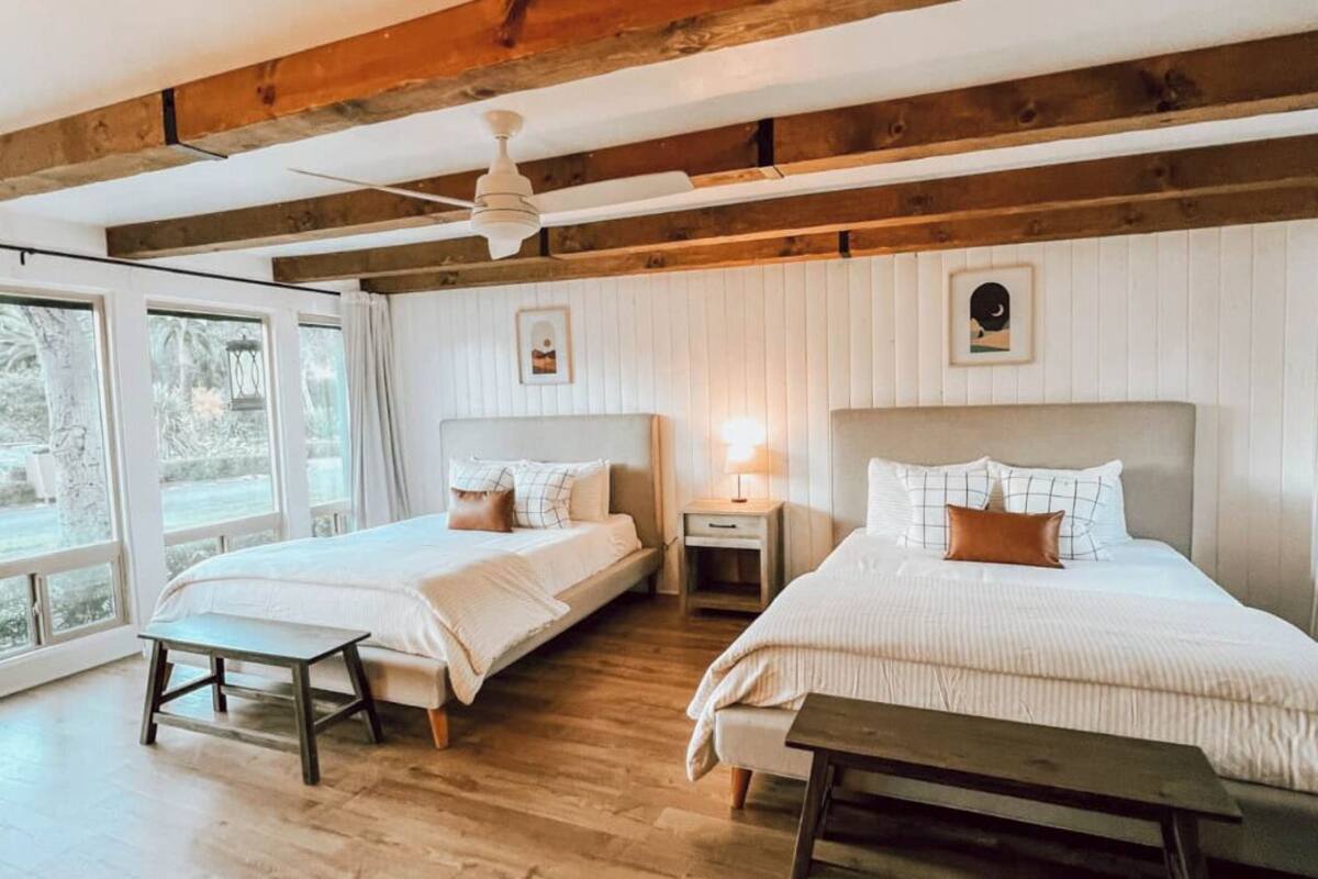 A bedroom with two beds, wooden beams, a ceiling fan, and large windows.