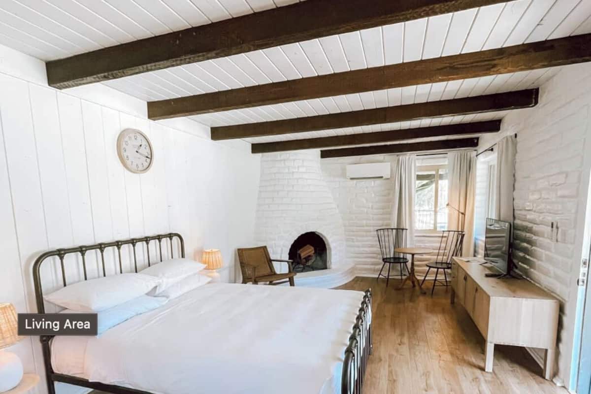 A bedroom with a wooden floor, a metal bed frame, a white wall clock, a fireplace, and a window with curtains.