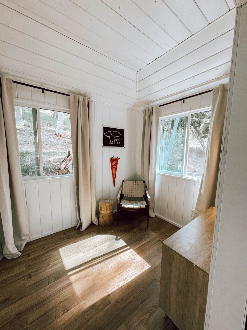 Une pièce aux murs et au sol en bois, avec une chaise, une petite table et une fenêtre donnant sur des arbres.