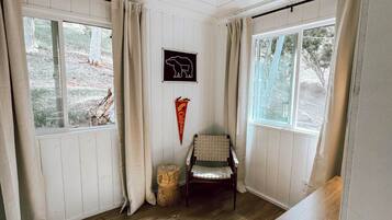 Une pièce aux murs et au sol en bois, avec une chaise, une petite table et une fenêtre donnant sur des arbres.