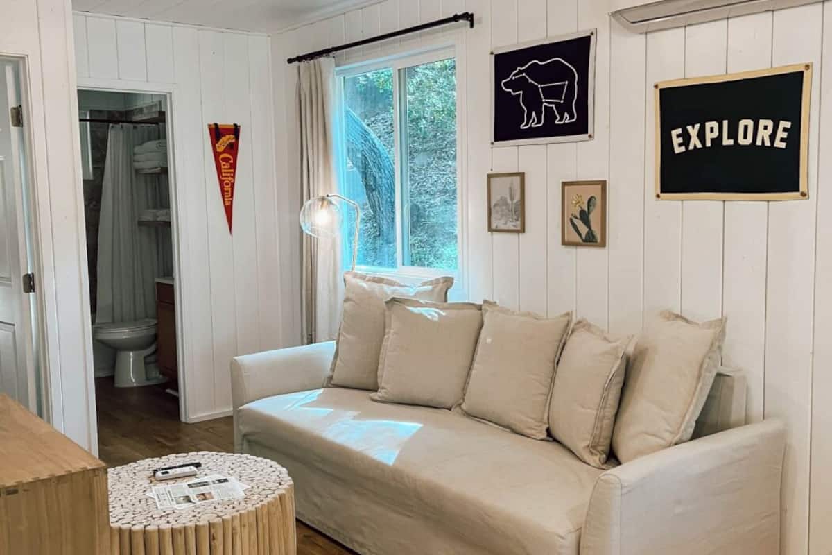 A cozy room with a beige sofa, wooden flooring, a round wooden table, and a window with a view of greenery.